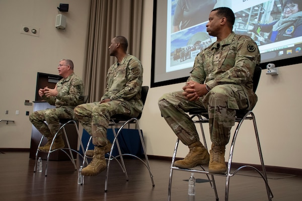 Three men wearing camouflage military uniforms sit on stools on a stage with a projection screen behind them.