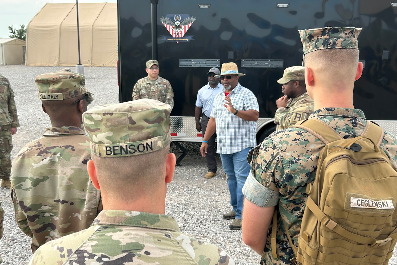 A man in civilian clothing talks to a group of people in camouflage military uniforms.