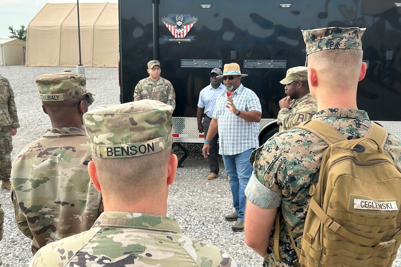 A man in civilian clothing talks to a group of people in camouflage military uniforms.