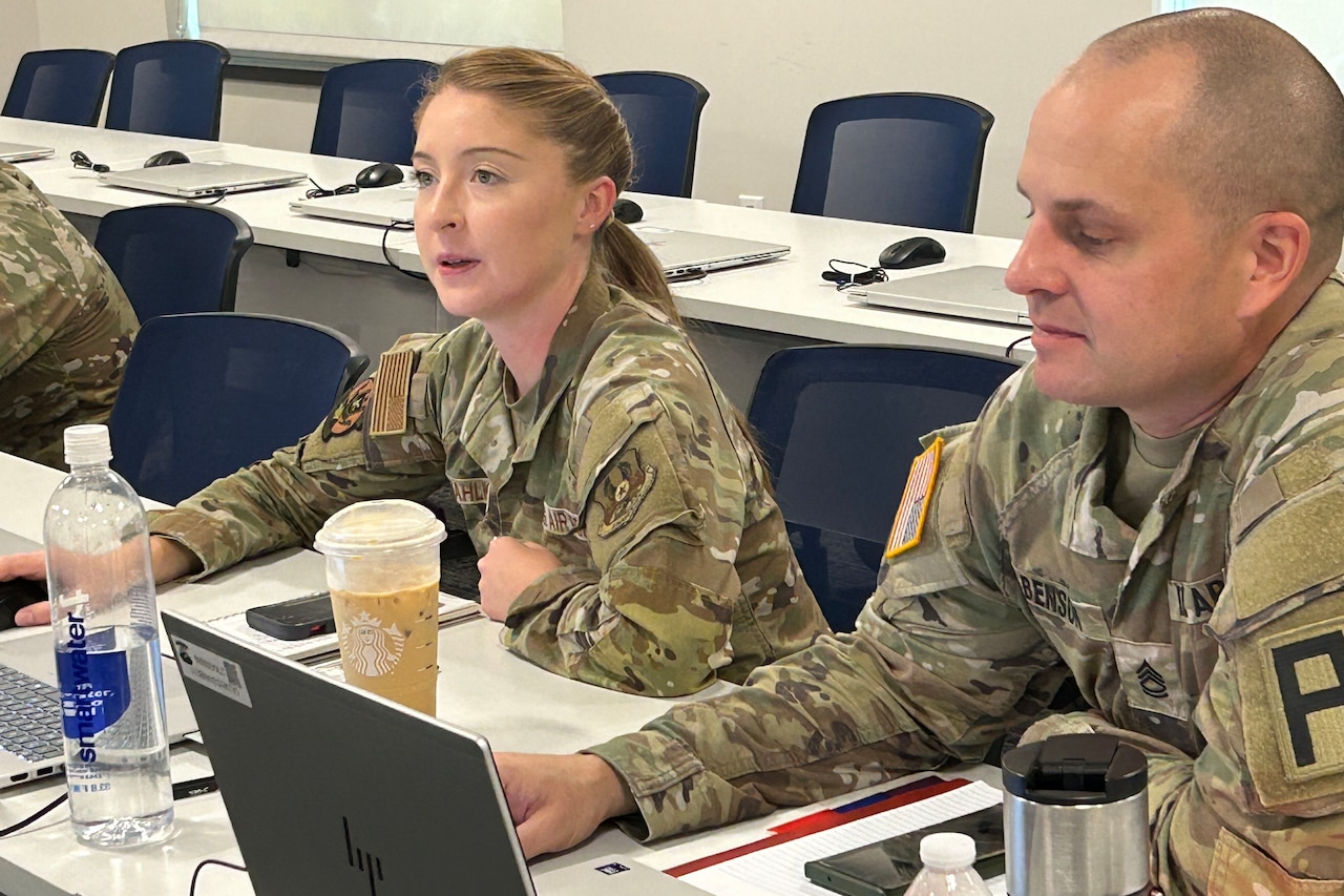 A man and woman in camouflage military uniforms look at laptop screens while sitting at a long desk.