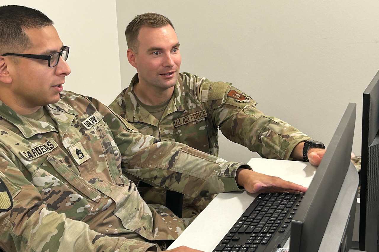 Two men wearing camouflage military uniforms sit at a computer and look at its monitor.