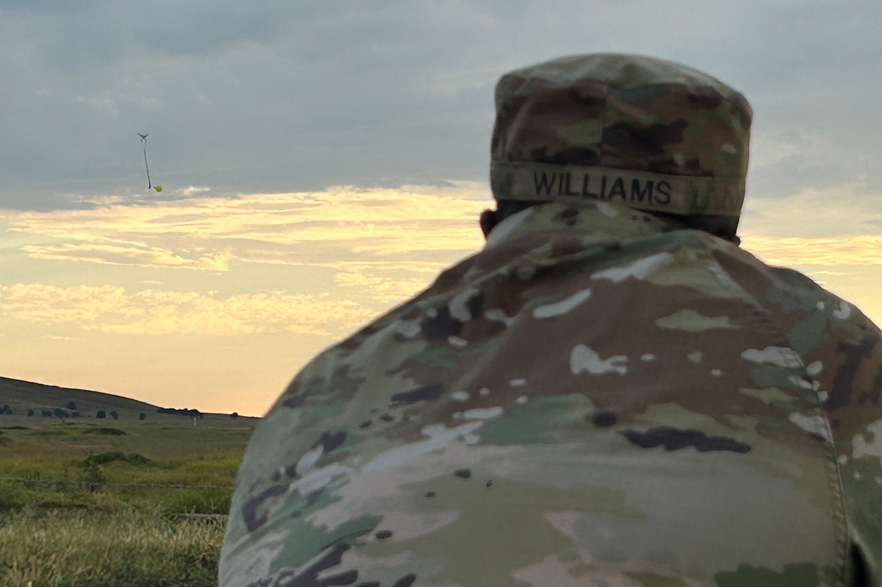 The back of a man wearing a military camouflage uniform is shown in the forefront while a drone, carrying a balloon, floats in the sky in the distance.