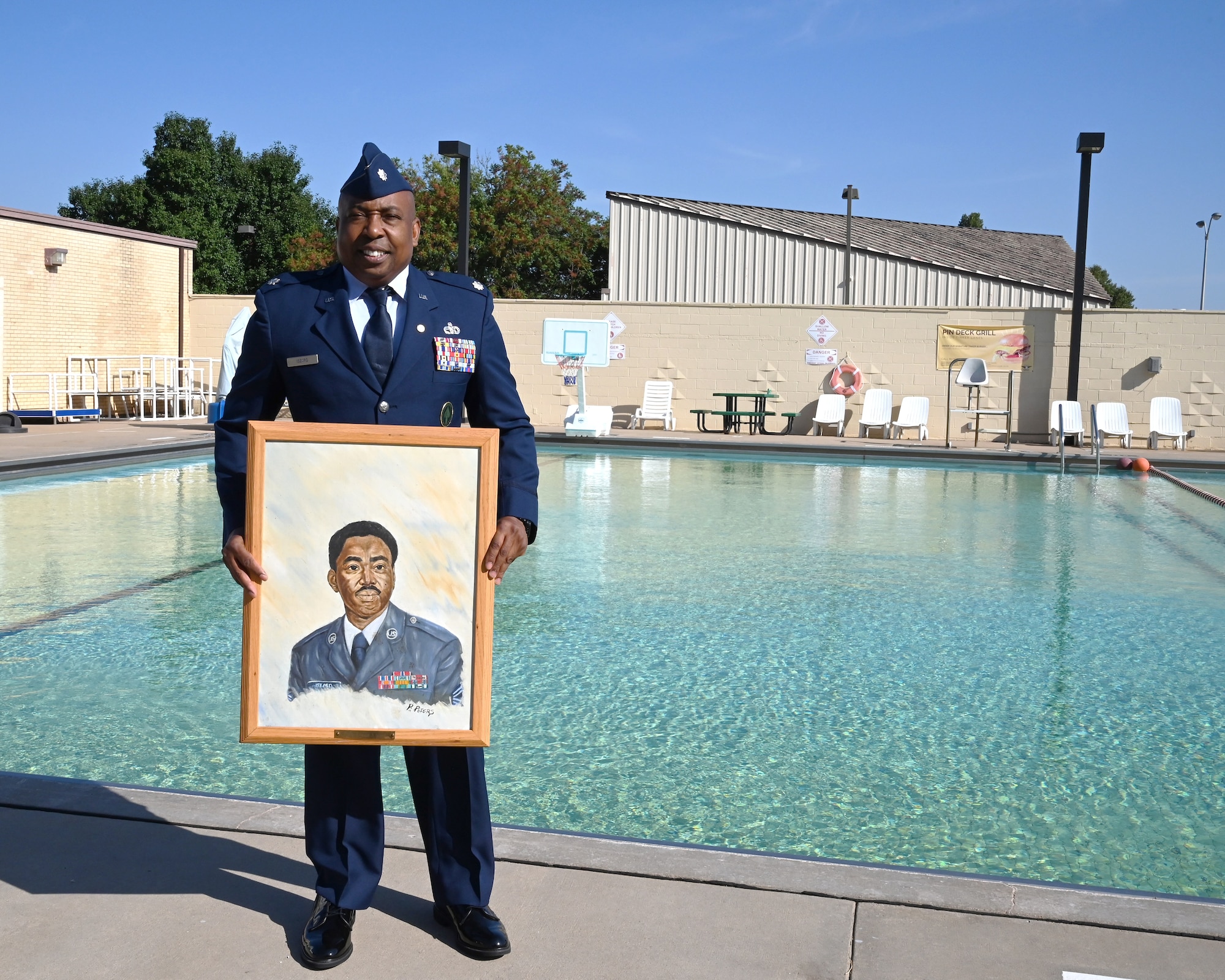 A man in dress blues stands at a pool, holding a painting of his uncle.