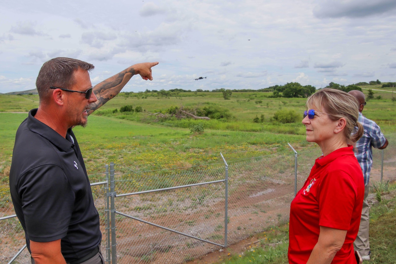 A man in business casual attire points toward a flying drone in a field as He talks to a woman in similar attire.