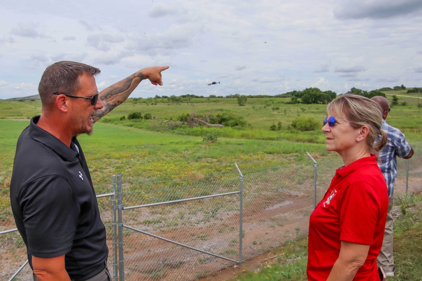 A man in business casual attire points toward a flying drone in a field as He talks to a woman in similar attire.