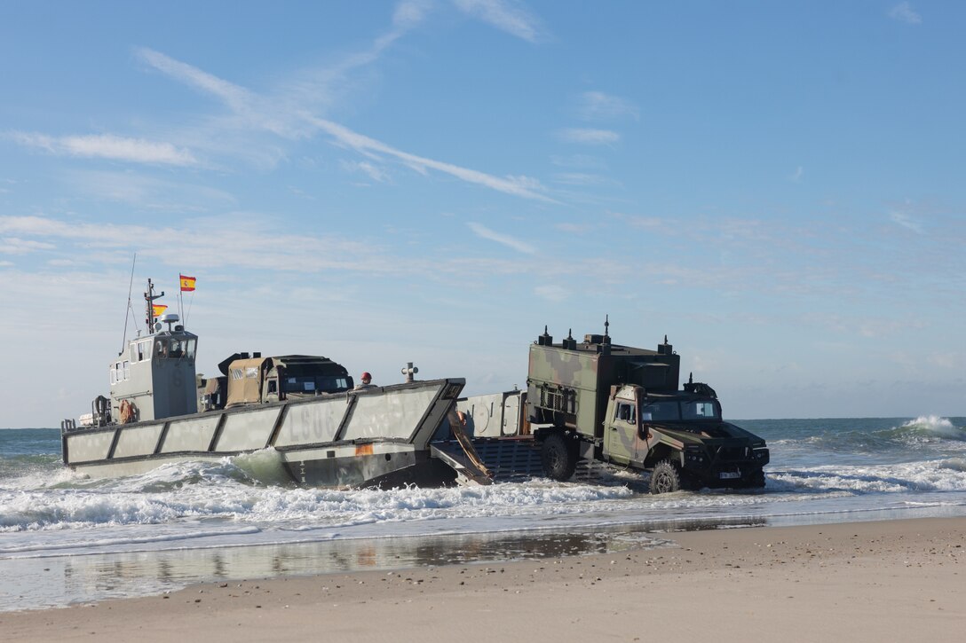 A military vehicle exits a water landing craft on a beach. The Spanish flag is flying from the landing craft and there is another vehicle waiting to exit.