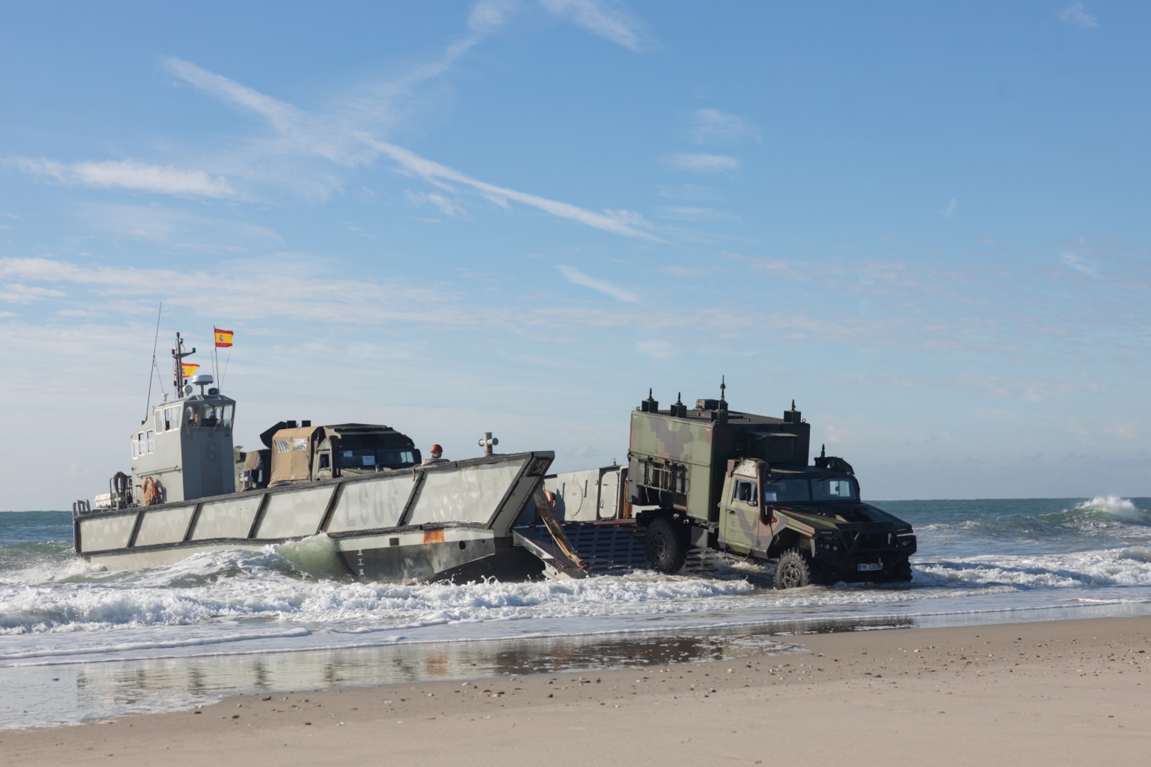 A military vehicle exits a water landing craft on a beach. The Spanish flag is flying from the landing craft and there is another vehicle waiting to exit.