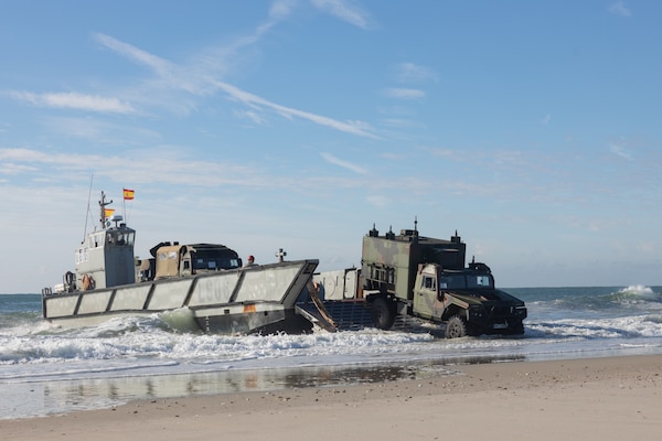 A military vehicle exits a water landing craft on a beach. The Spanish flag is flying from the landing craft and there is another vehicle waiting to exit.