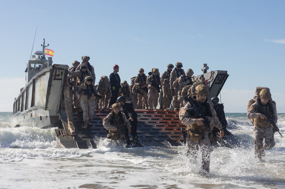 Dozens of people wearing military camouflage uniforms and holding rifles depart a water landing craft as waves crash around them on a beach. A Spanish flag is flying from the landing craft in the background.