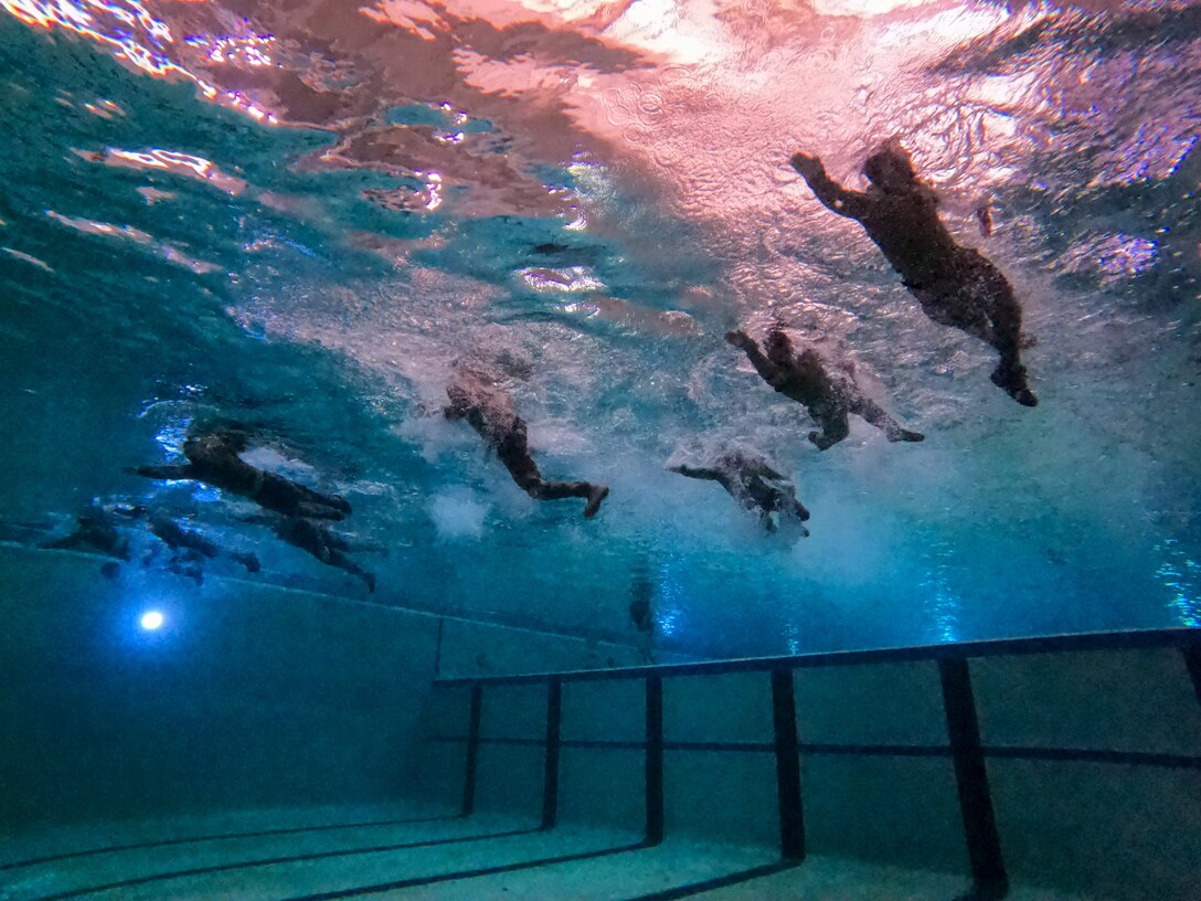 Eight people wearing camouflage military uniforms swim in a pool during water survival training. The photo is taken from the bottom of the pool looking up at the people swimming on the surface.