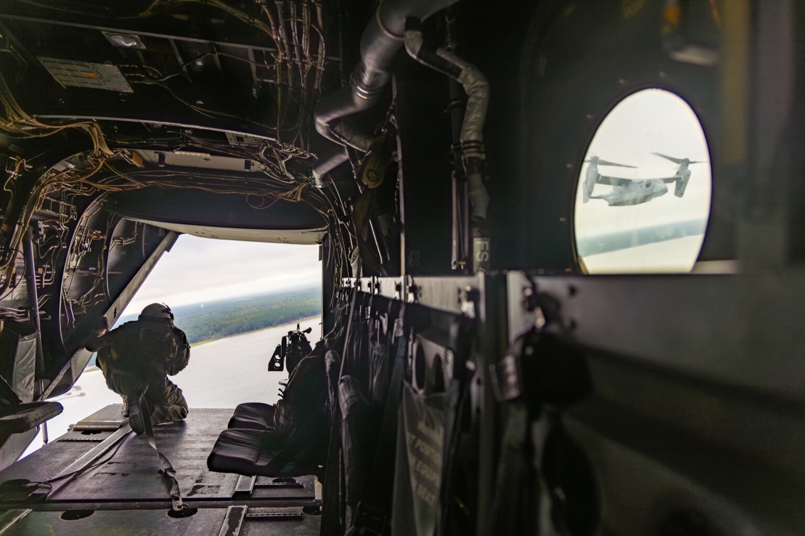 A person wearing a flight suit and helmet looks out the back of an open military aircraft. There is a strap hooked to the floor of the aircraft that is also attached to the person for safety purposes. You can see exposed wires, seats and other equipment in the aircraft. Another military aircraft is visible from a window on the right.