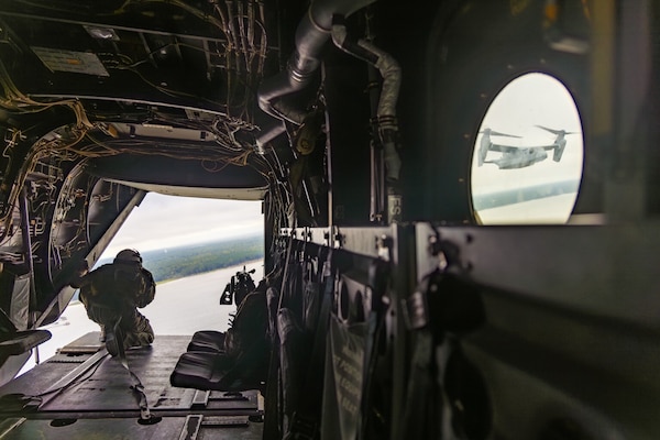 A person wearing a flight suit and helmet looks out the back of an open military aircraft. There is a strap hooked to the floor of the aircraft that is also attached to the person for safety purposes. You can see exposed wires, seats and other equipment in the aircraft. Another military aircraft is visible from a window on the right.