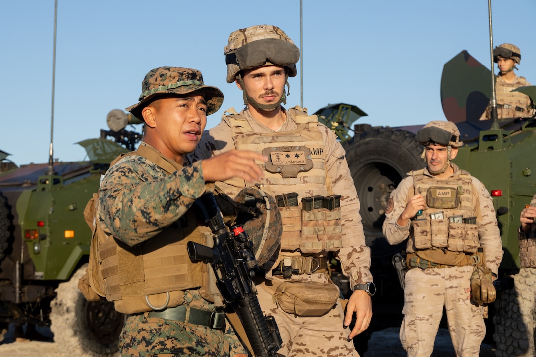 A person wearing a military camouflage uniform speaks to a foreign military member in similar attire while gesturing with his right hand. There are two other foreign military members in similar attire standing in front and on top of a military vehicle.