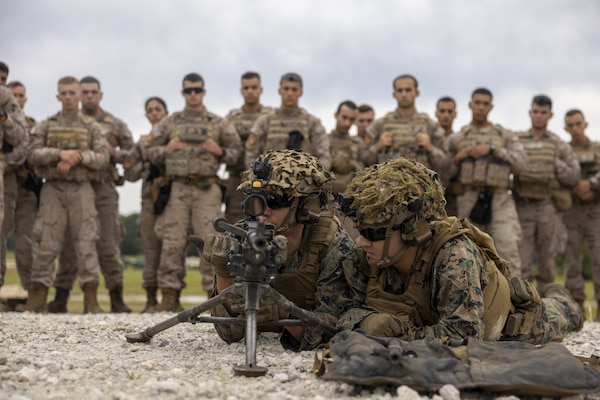 A Marine wearing a camouflage military uniform, helmet and sunglasses lies on the ground as he looks through the scope of a machine gun. Another man in similar attire lies next to him while approximately a dozen foreign military members in camouflage uniforms observe in the background.