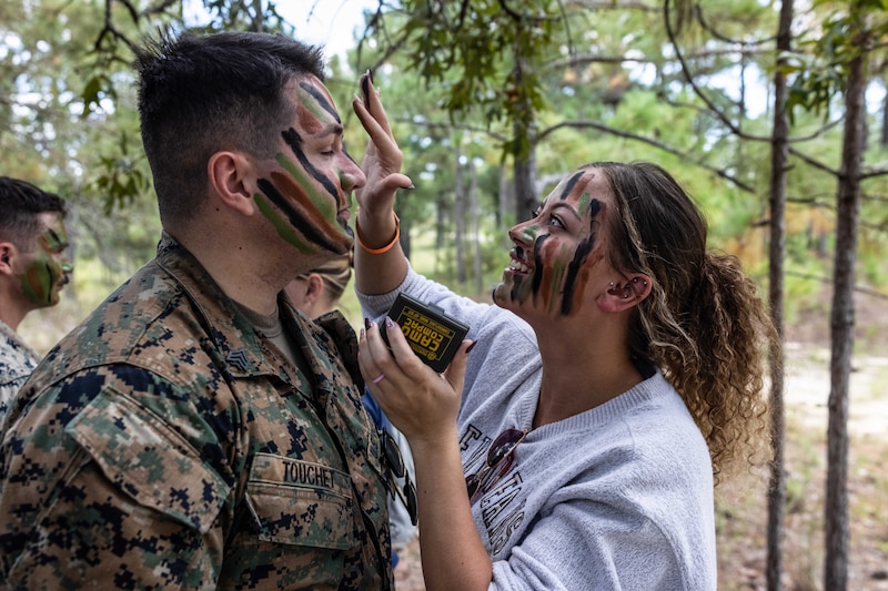 A Marine's wife applies camouflage paint to their face in a wooded area.