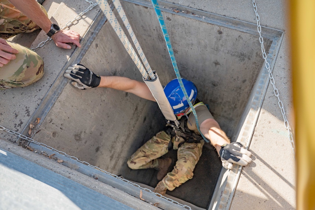 An airman harnessed to a pulley system sits in a square crawlspace.