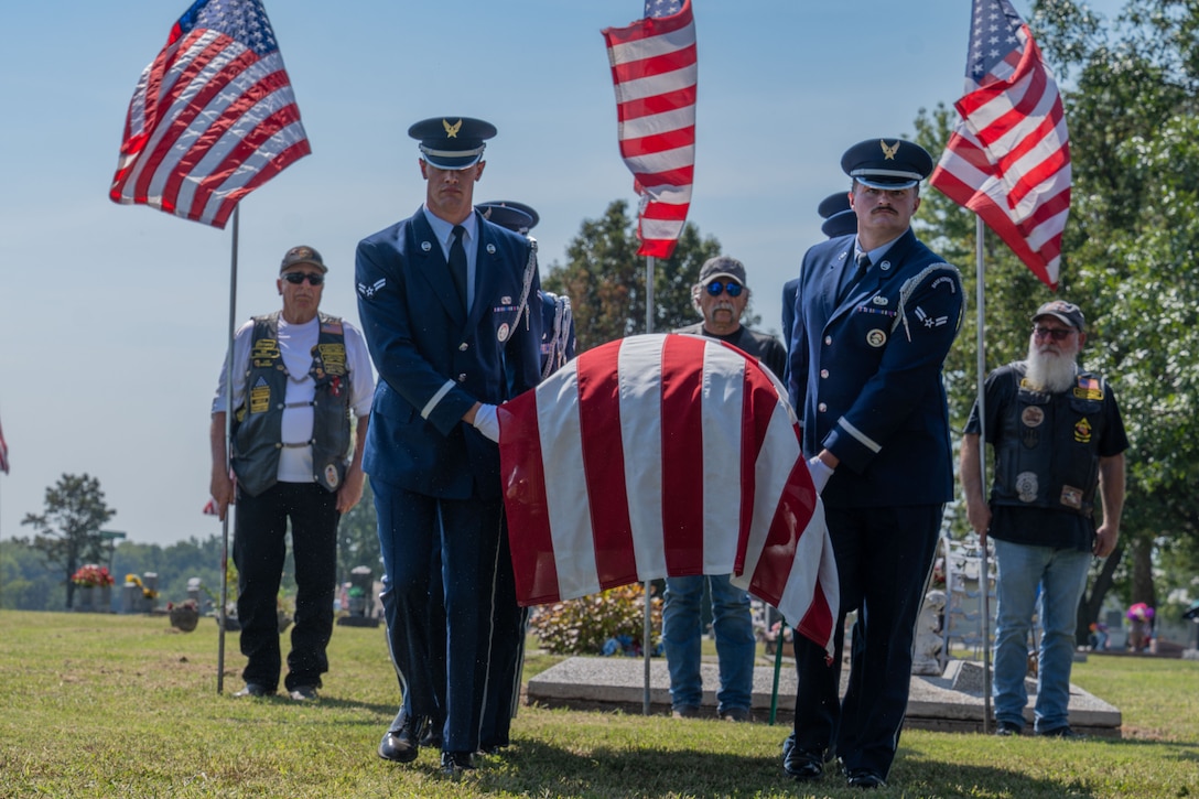 Service members in ceremonial dress carry a casket draped with an American flag at a burial site as civilians hold flags in the background.