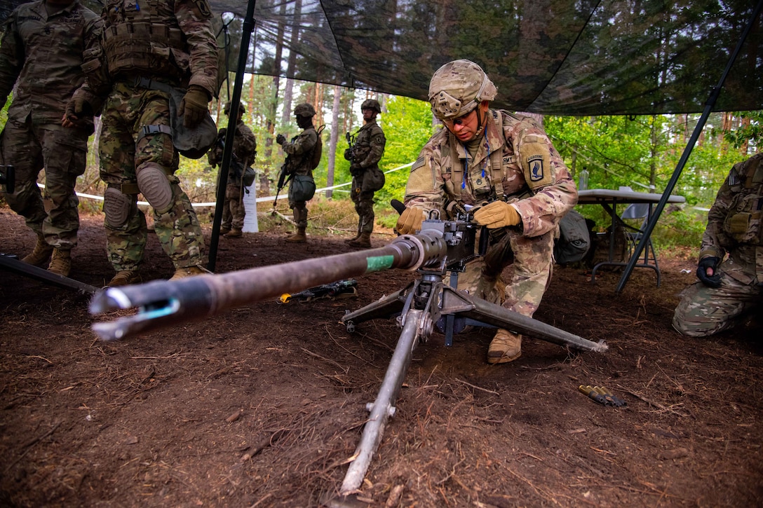 A soldier kneels in dirt to aim a machine gun while other soldiers stand around under a camouflage tarp in a forest setting.
