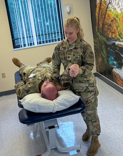 an airman lays on a physical therapy table while another airman assists him with stretching his shoulder