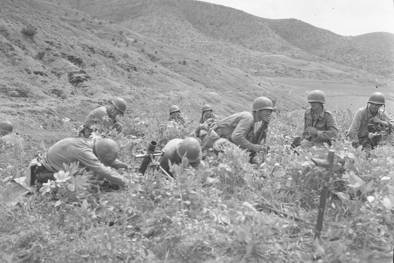 Nearly a dozen people in combat uniforms and helmets huddle in tall grass.