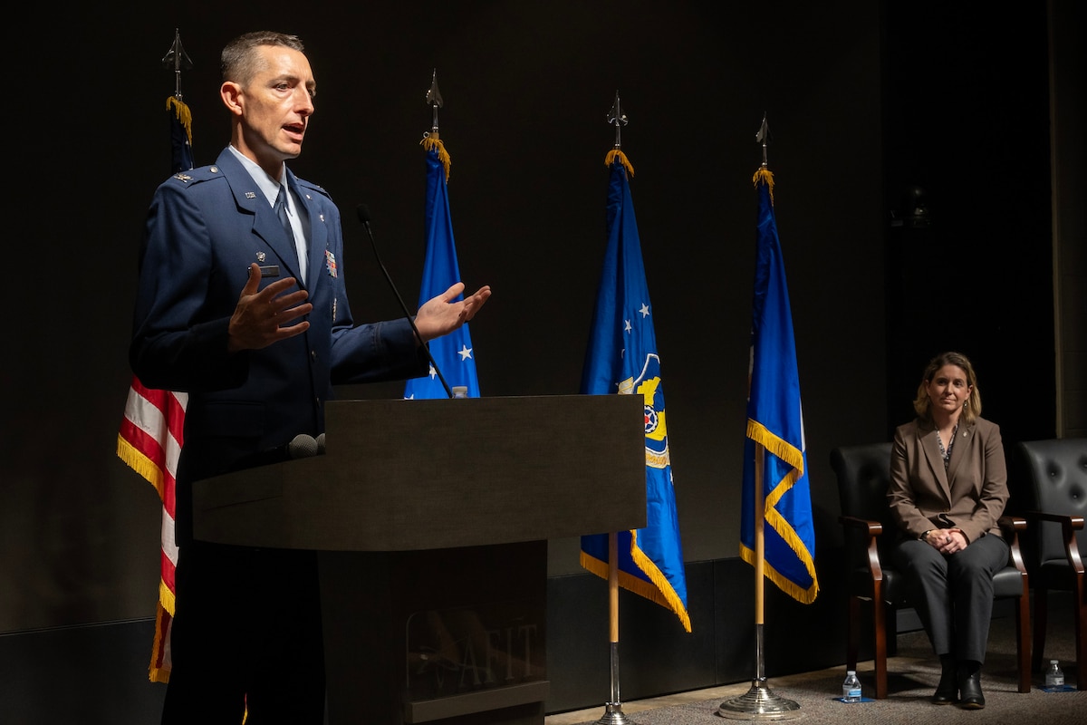 Man in Air Force blues stands behind podium. Woman in brown jacket sits on chair.