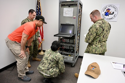 A group of U.S. Navy sailors and instructors work on electronic equipment in a lab setting. Wires and circuit boards are visible.