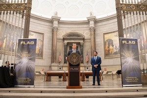 A man in business attire is standing behind a podium delivering remarks in a large, circular ornate room with marble walls. There are two American flags behind him, and another man off to the side observing him. On either side of the podium are banners that read “FLAG - Two Million Milestone.”