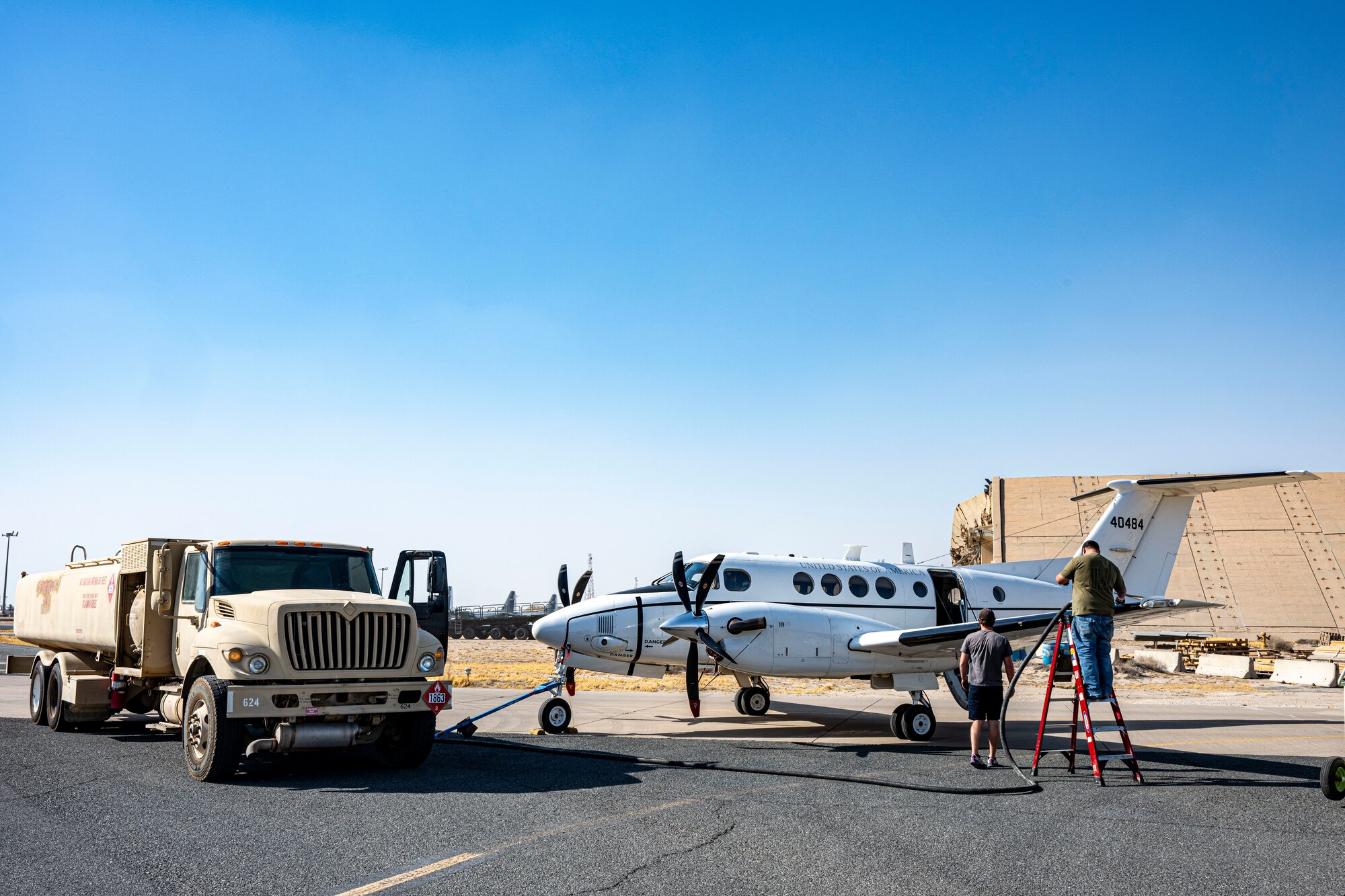 A C-12 Huron sits right of frame facing left of frame toward a refueler vehicle as contractors prepare to fuel the aircraft