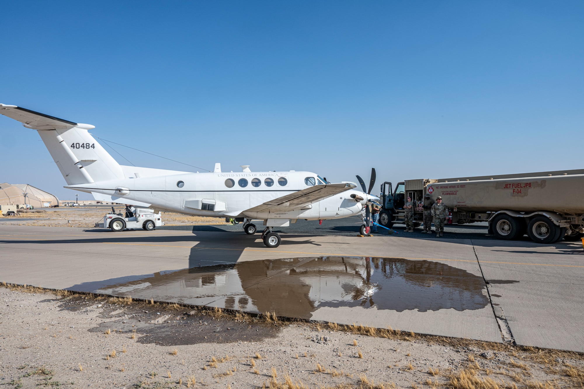 A C-12 Huron sits left of frame next to a refueler truck right of frame, both in the background, while a puddle of simulated fuel sits in the foreground