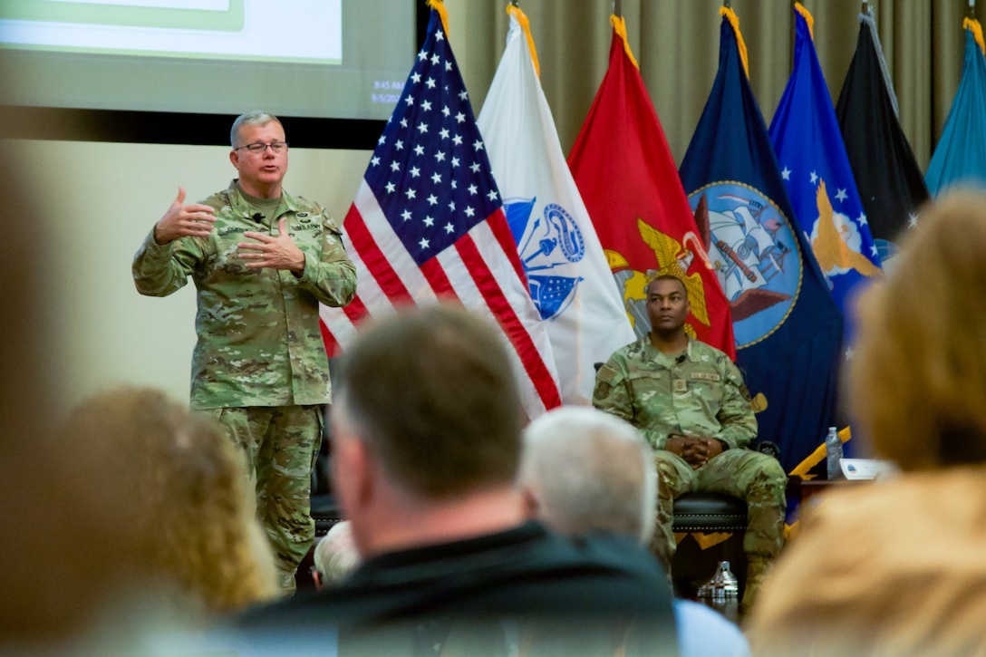 A man wearing a camouflage military uniform stands on a stage addressing a large audience.