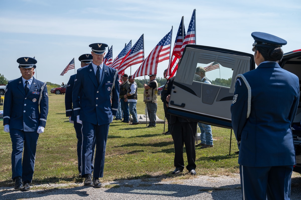 A group of people in military dress uniforms march toward a hearse.