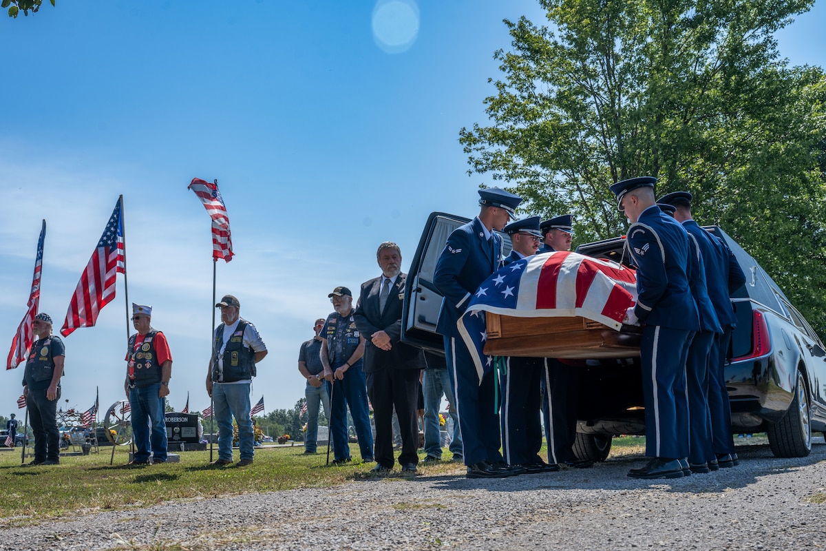 People in military dress uniforms pull a casket draped in an American flag out of a hearse.
