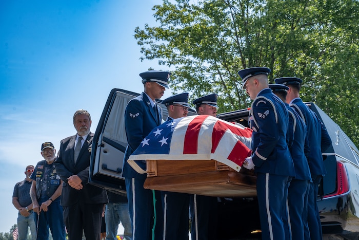 People in military dress uniforms pull a casket draped in an American flag out of a hearse.