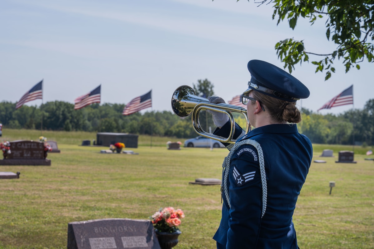 A person in a military dress uniform holds a bugle to their mouth at a cemetery.