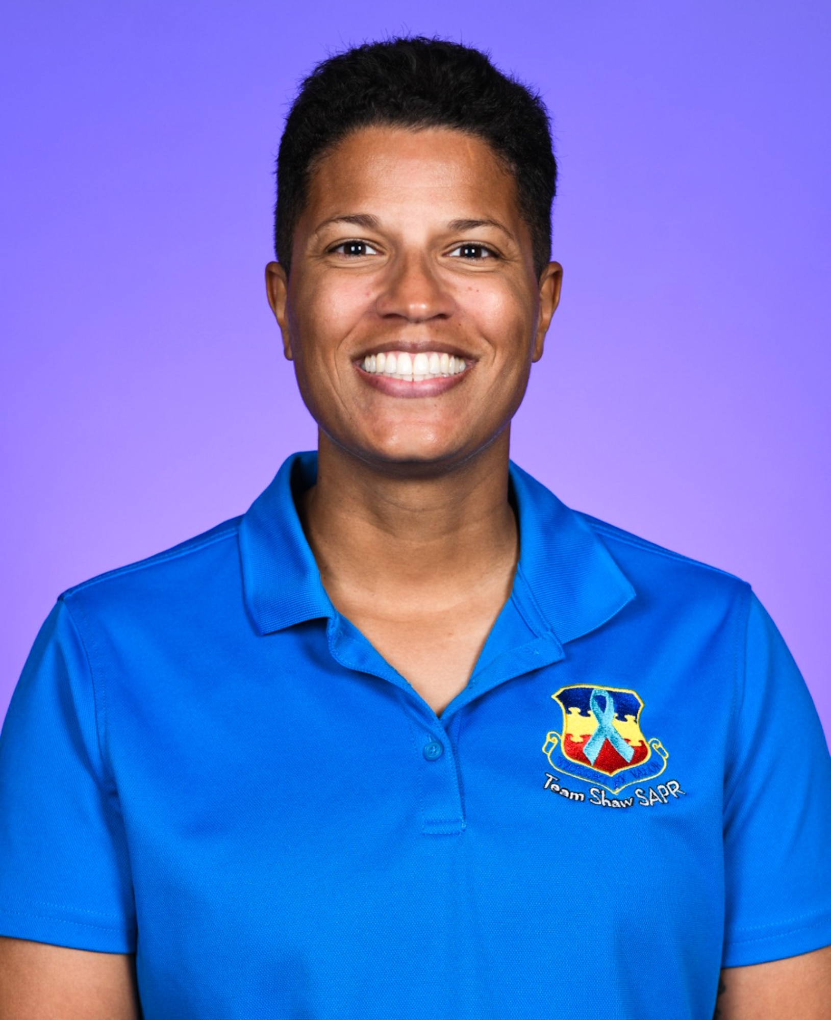 A woman poses, smiling, for a photo in a blue shirt. She stands in front of a purple background.