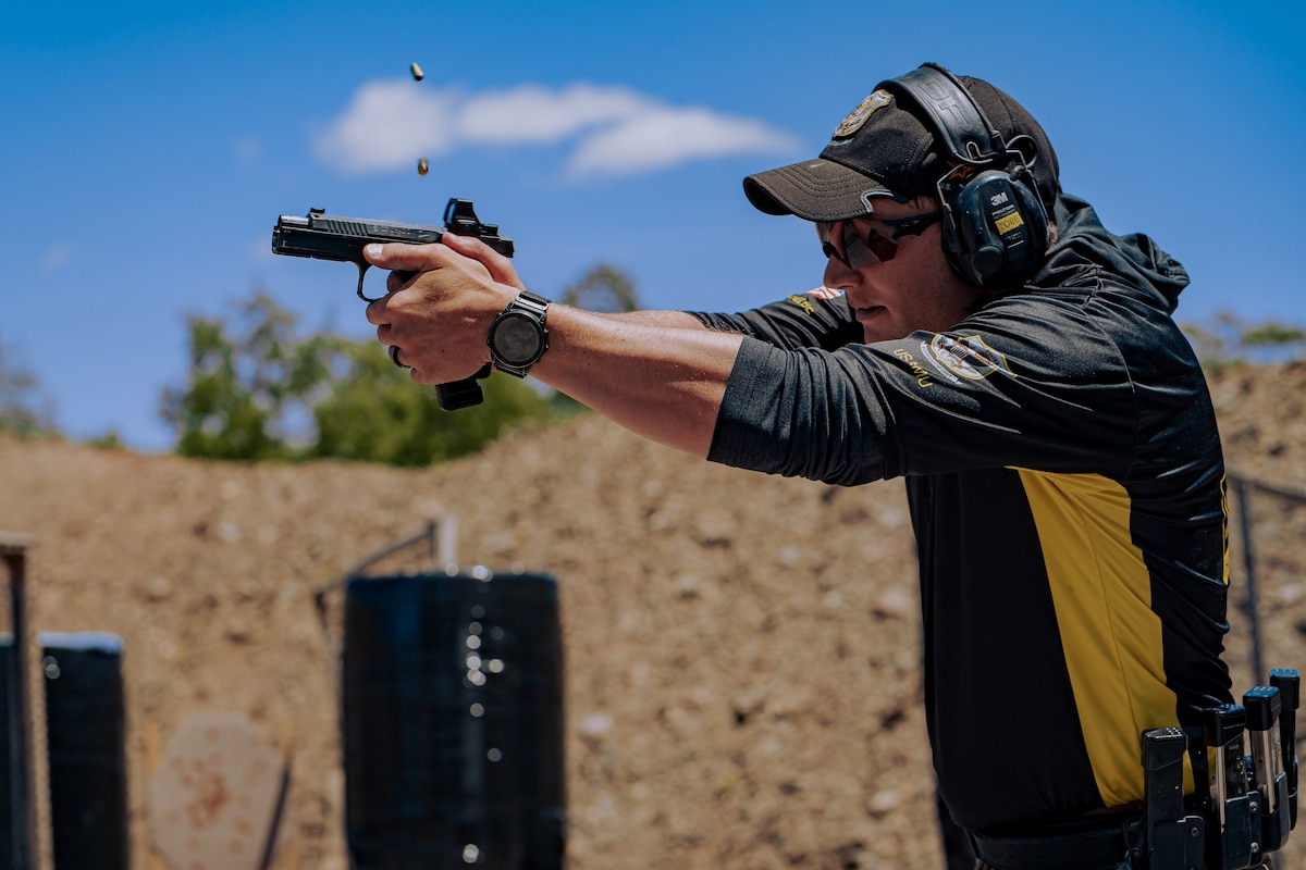 A man wearing a hooded shirt, hat and hearing protection fires a handgun in front of a brick wall. Bullet casings fly out of the gun in the air.