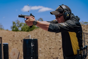 A man wearing a hooded shirt, hat and hearing protection fires a handgun in front of a brick wall. Bullet casings fly out of the gun in the air.