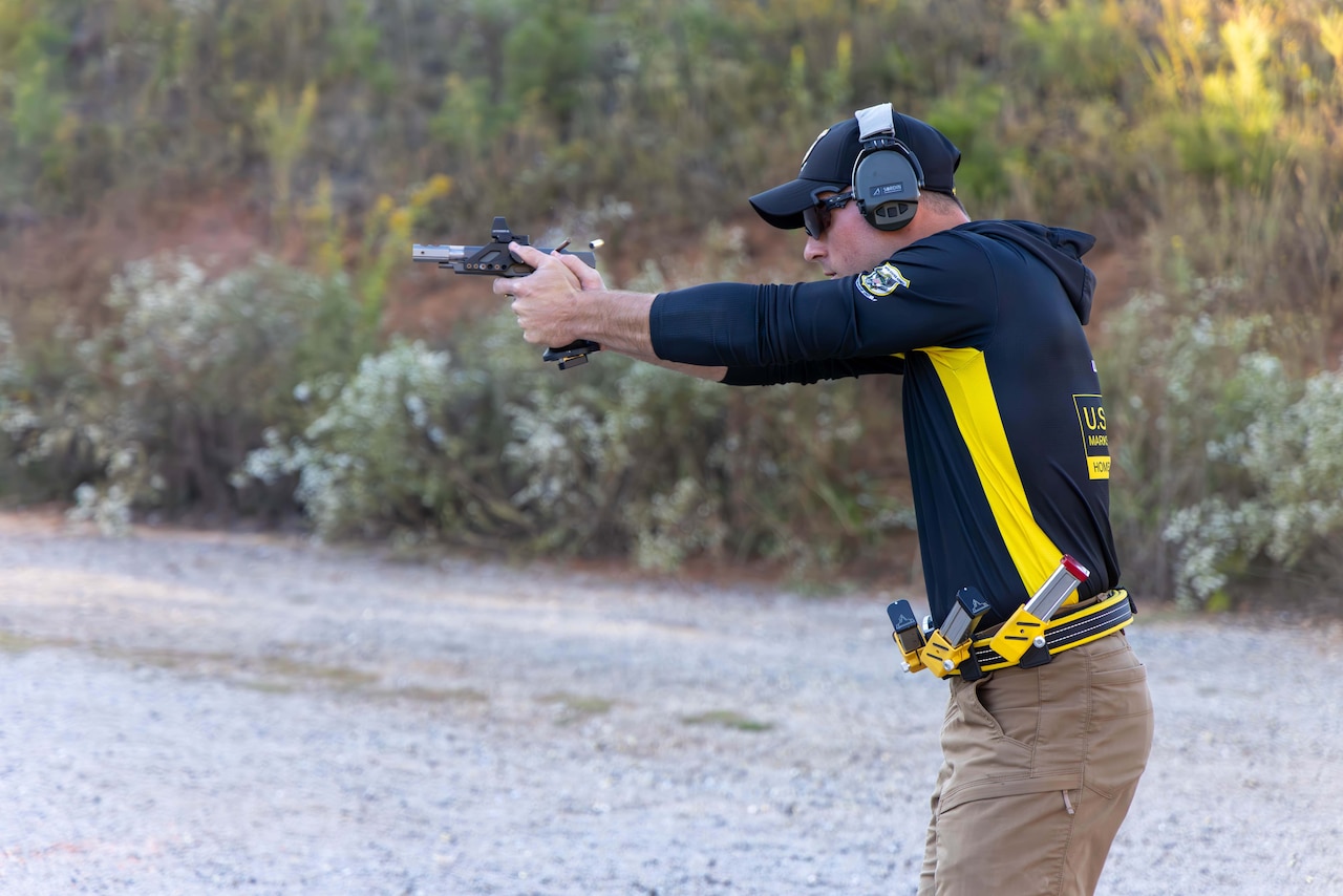 A man wearing a hooded shirt, hat, hearing protection and a belt carrying three magazines fires a handgun in front of a brush hill. A bullet casing flies out of the gun in the air.