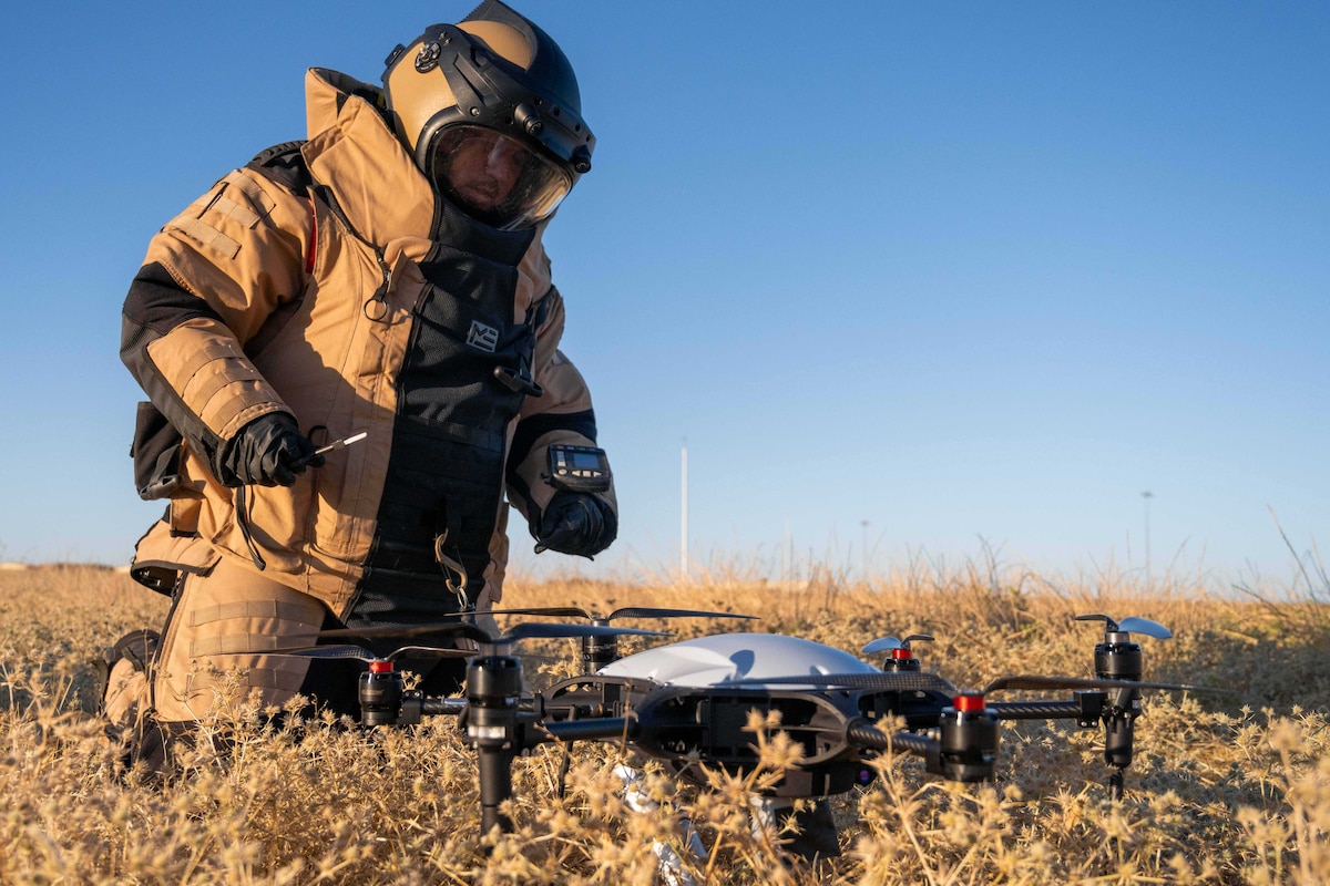 In a field, a person in a bomb suit kneels in front of a drone. The person is holding a tool in their right hand.