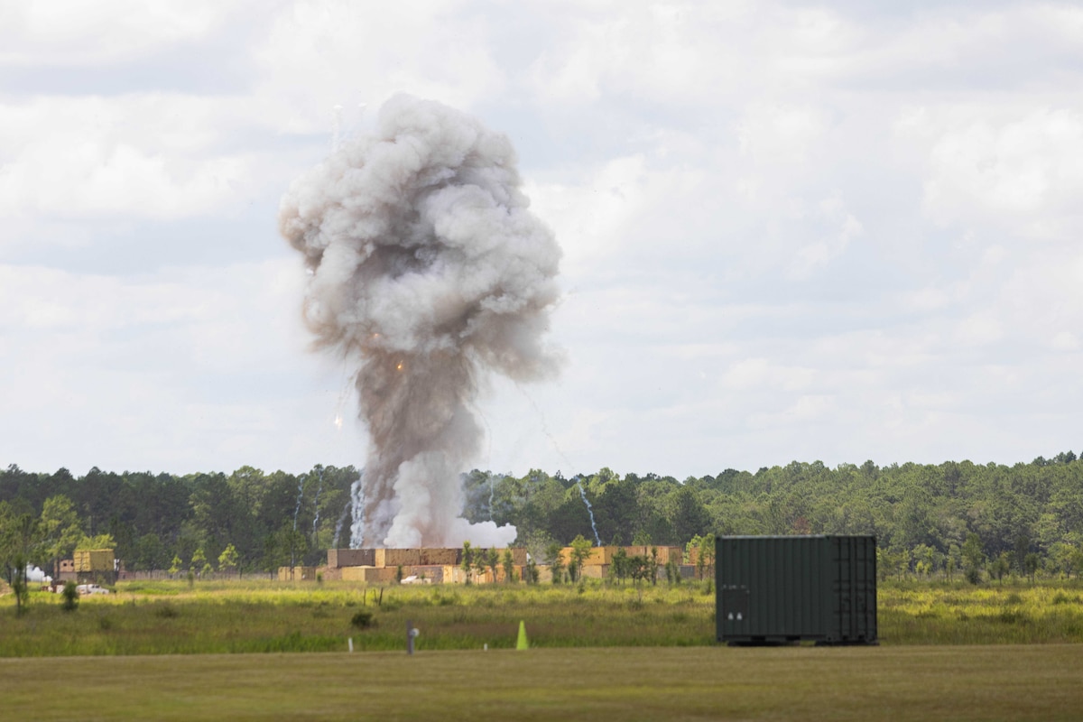 A plume of gray smoke rises from an explosion in the distance. There are trees in the background and shipping containers surrounding the area where the explosion occurred.