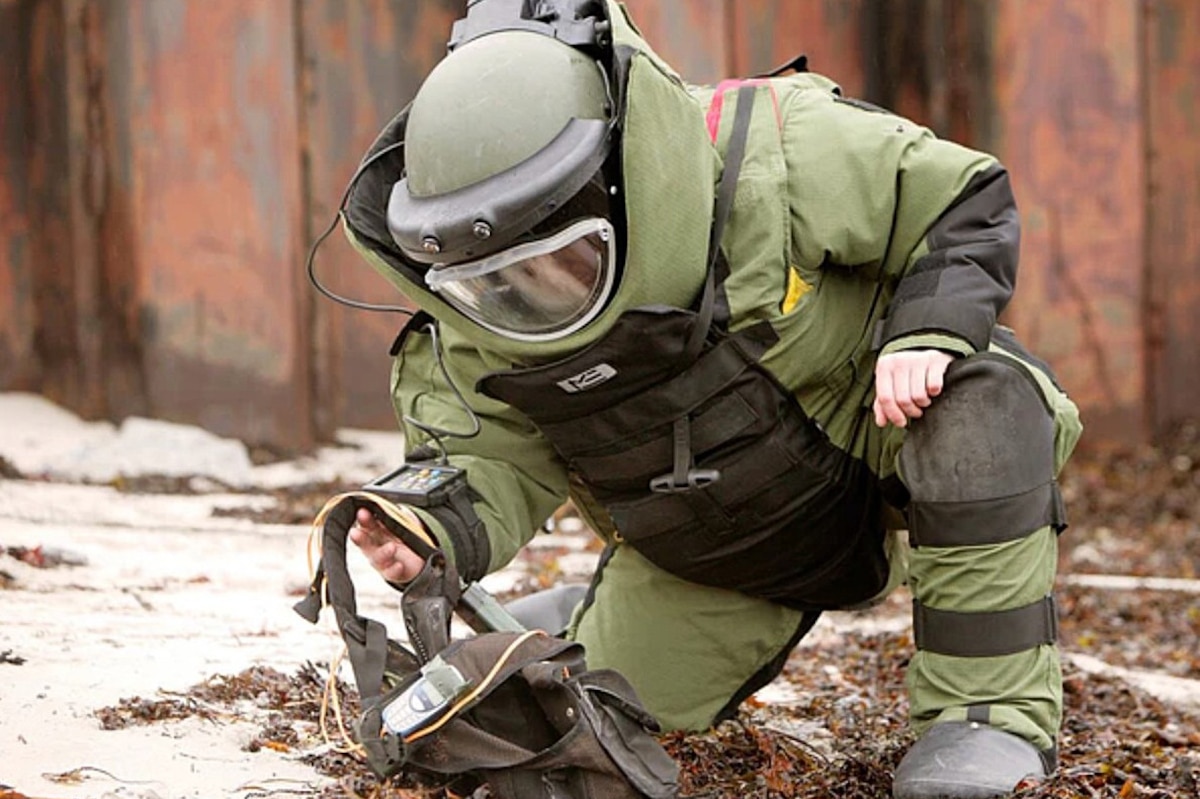 A person in a bomb suit kneels down to pick up a bag lying on the ground.