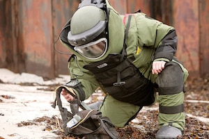 A person in a bomb suit kneels down to pick up a bag lying on the ground.