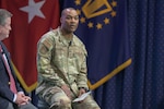 A man in an Air Force uniform is seated on a stage with military flags behind him.