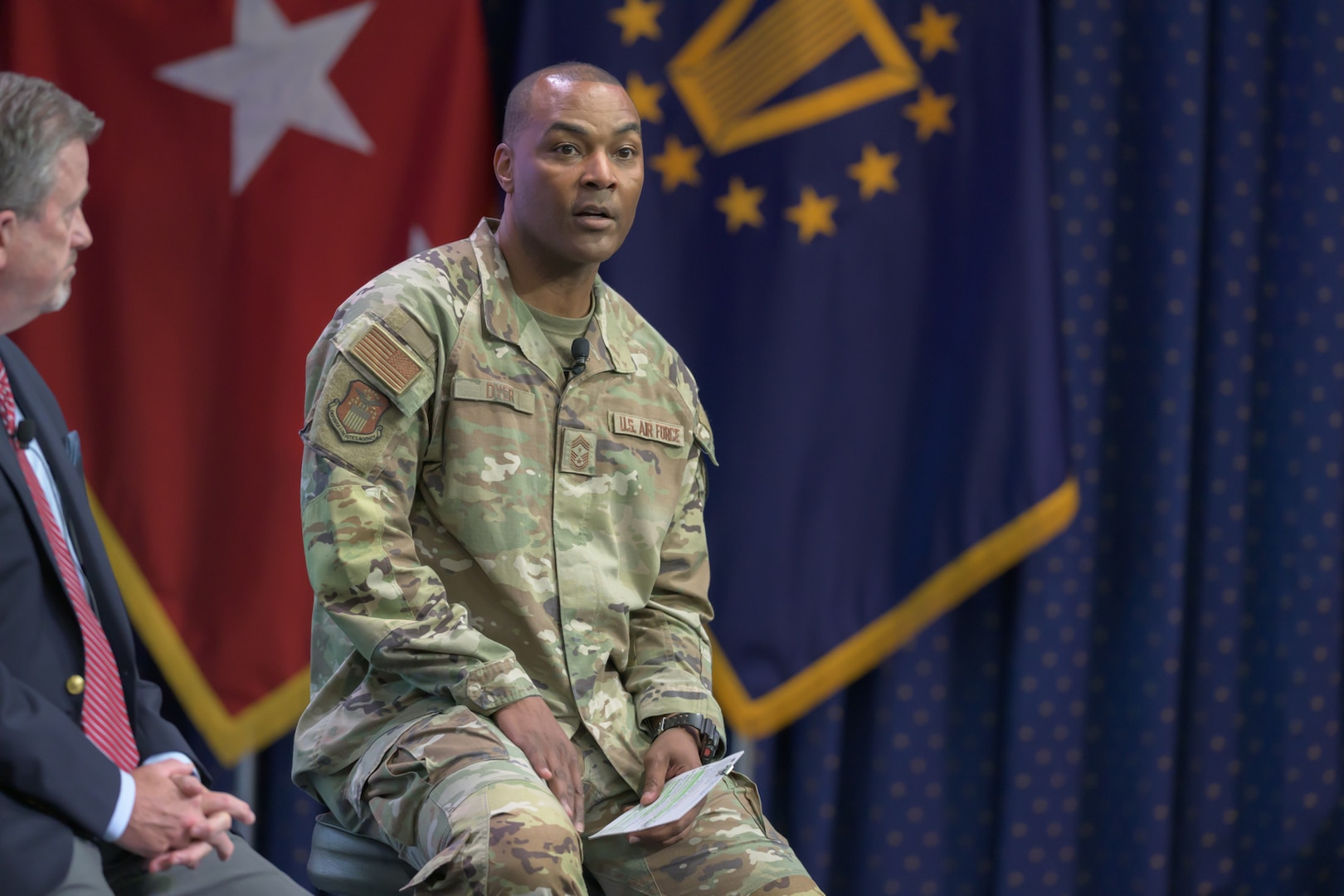 A man in an Air Force uniform is seated on a stage with military flags behind him.