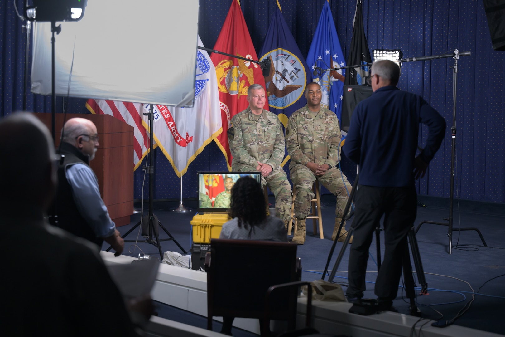 Two men sit on stools while a film crew is preparing to record them.