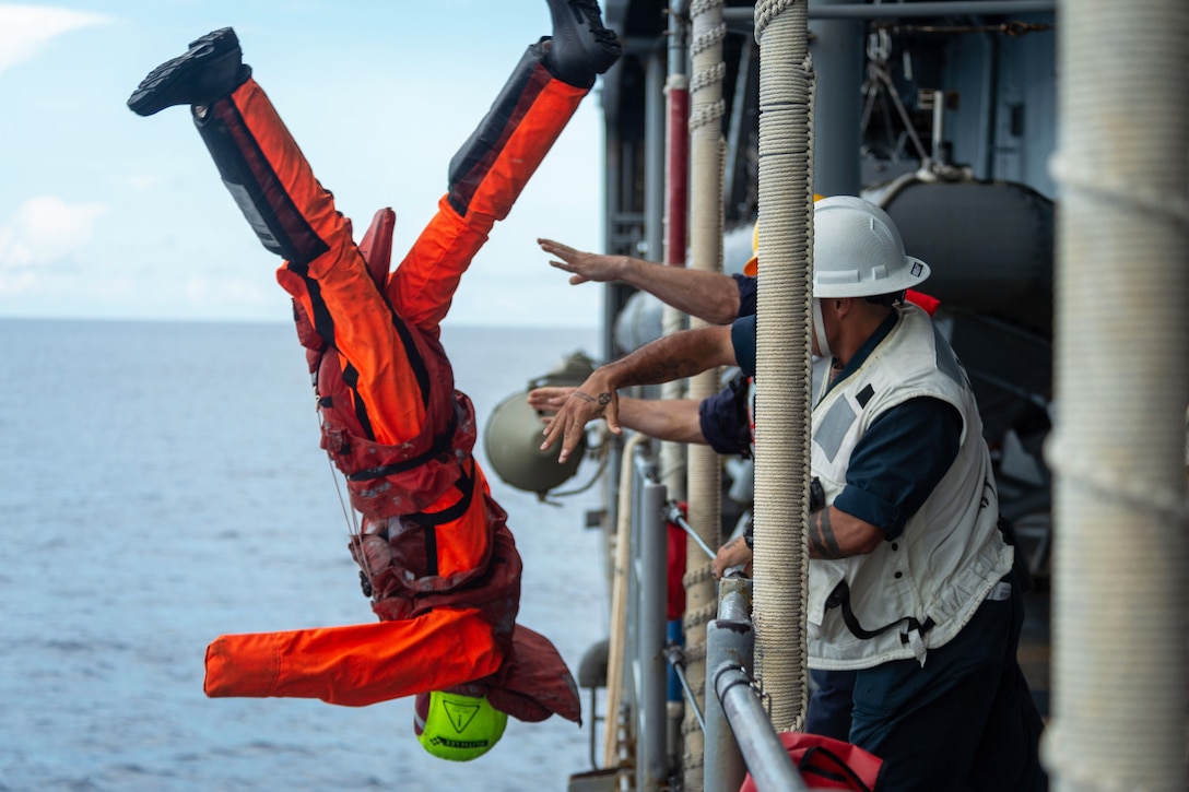 Sailors toss a mannequin wearing red gear off the side of ship as it transits a body of water during the day.