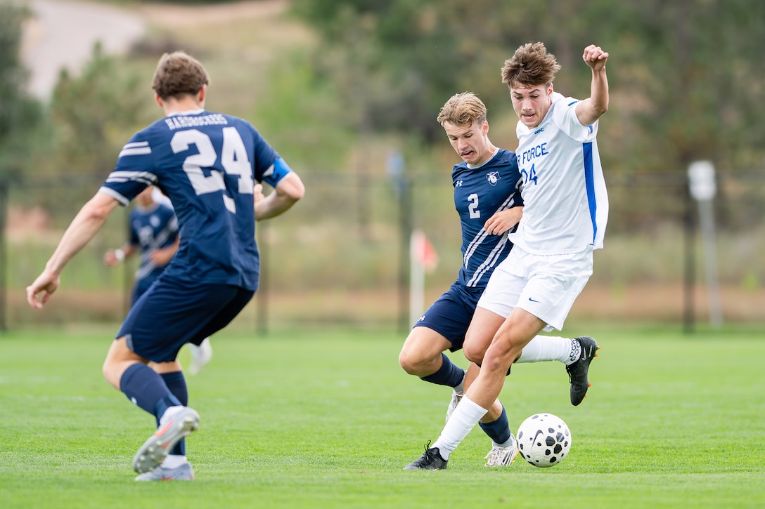 Two soccer players go after a ball during a match on a turf field in daylight.