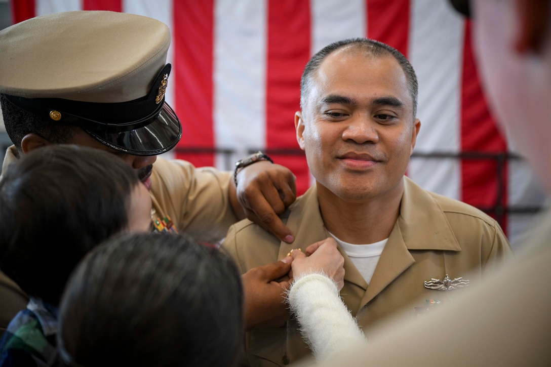 A close-up of a family adding a pin to a sailor’s lapel with an American flag in the background.