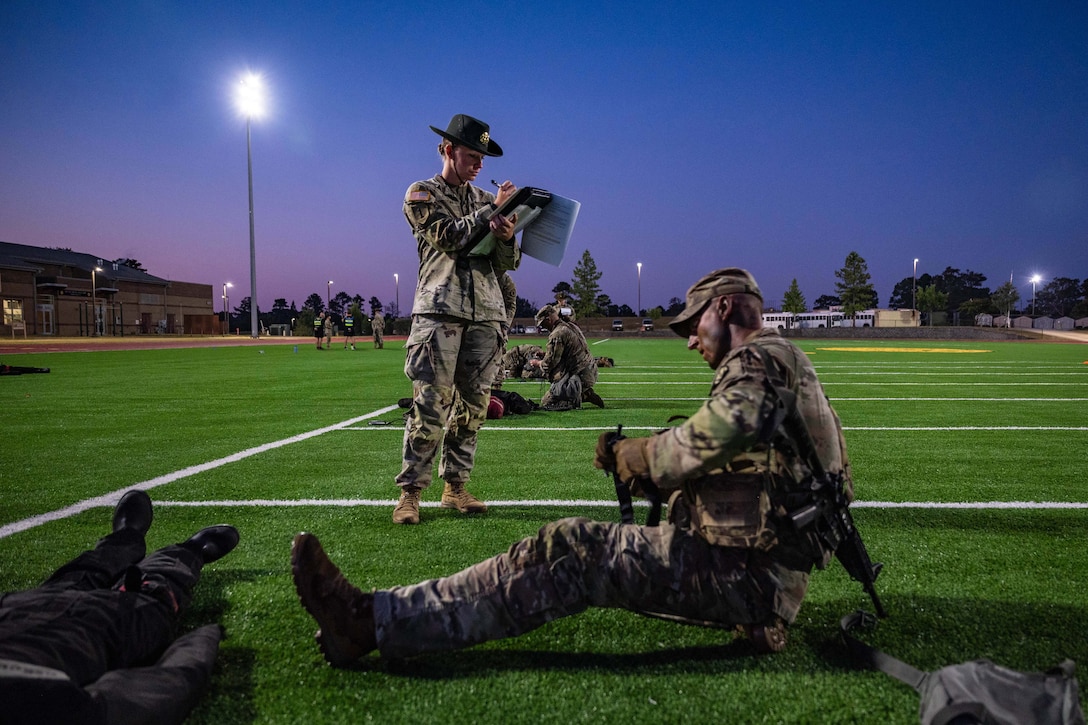 A soldier writes on a board as soldiers compete on a turf field in the dark illuminated by stadium lights.