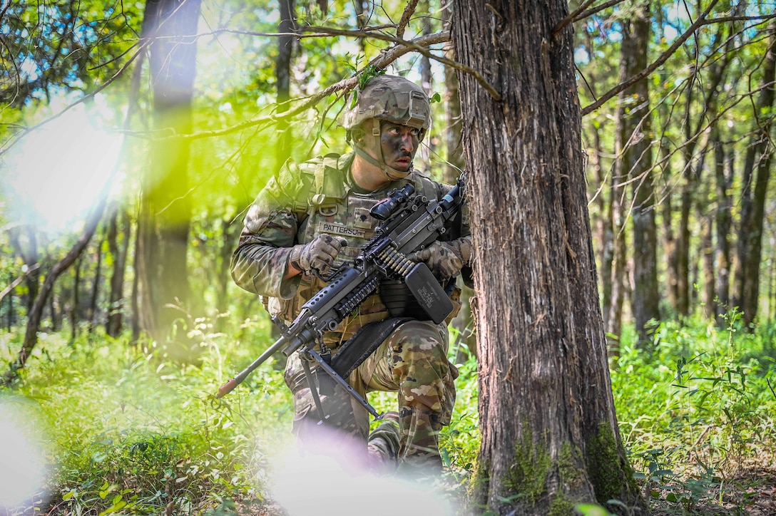 A National Guardsmen carrying a weapon kneels behind a tree in the woods during the day.